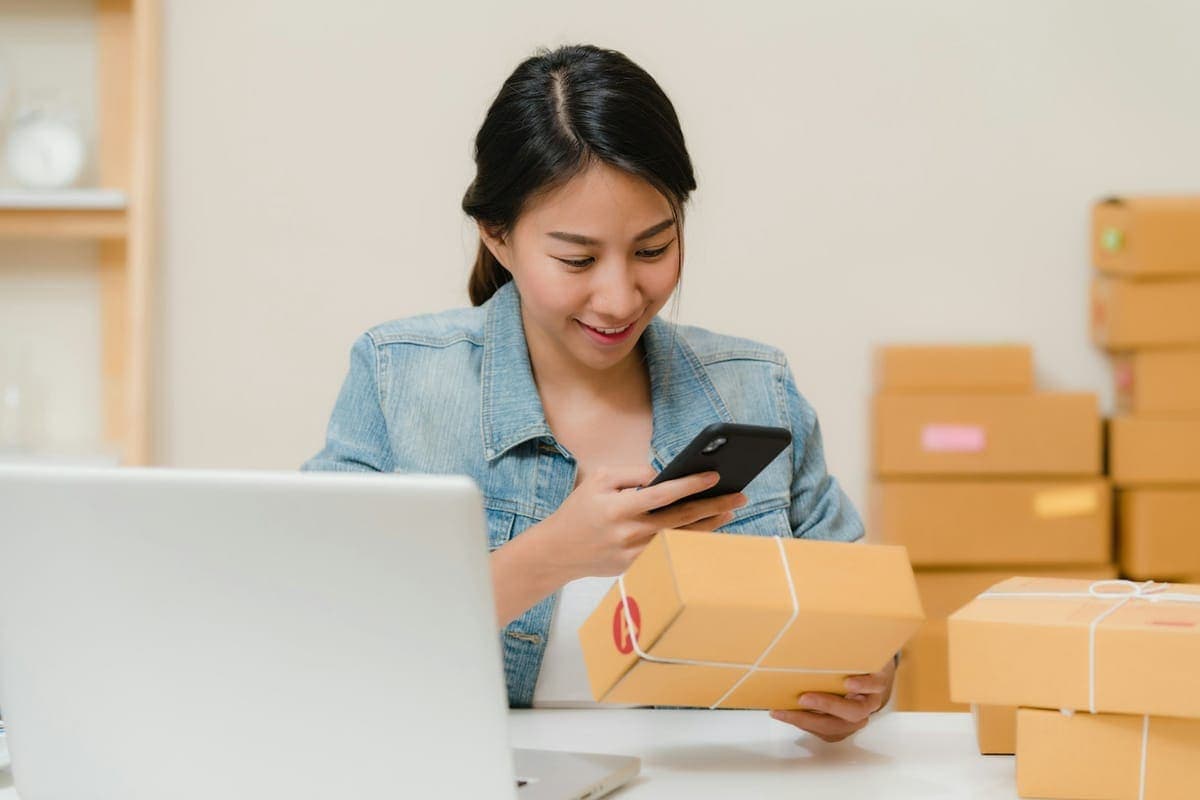 A founder checks an order on her phone while packing parcels at her laptop